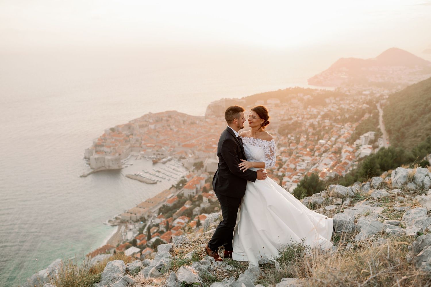 A couple in wedding attire embraces on a cliff overlooking the Adriatic coastline near Dubrovnik during golden hour.