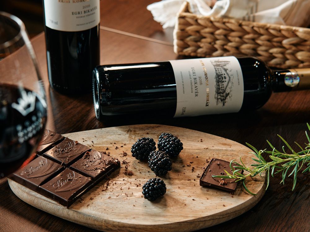 Dark chocolate bar with blackberries and rosemary on wooden board next to wine bottles.
