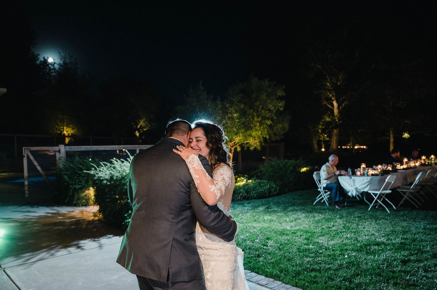 Couple sharing their first dance at an outdoor evening wedding reception with tables and lights in the background.