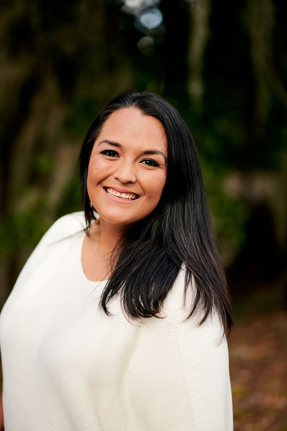 Portrait of a woman with long dark hair wearing a white sweater outdoors.