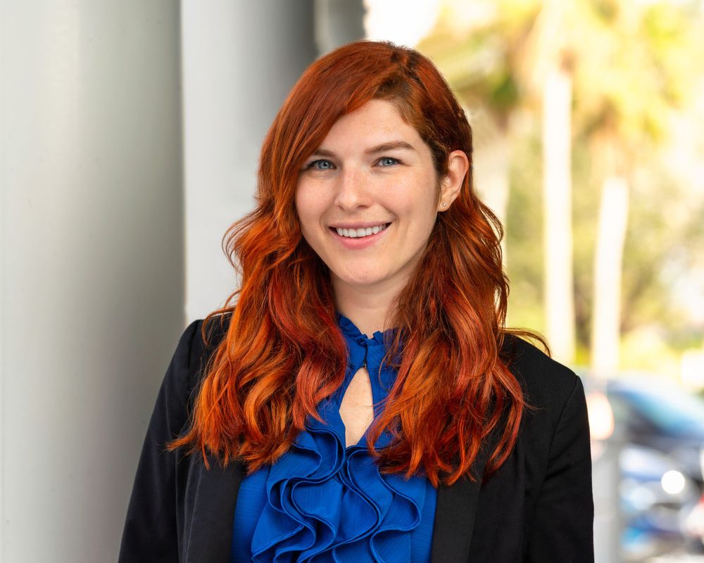 Professional portrait with vibrant red hair and blue blouse against bright outdoor background.