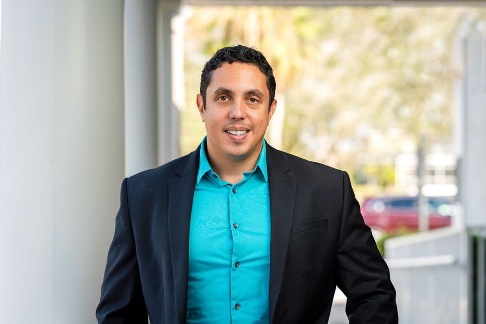 Professional wearing a black suit jacket and turquoise dress shirt smiling at camera in office setting.
