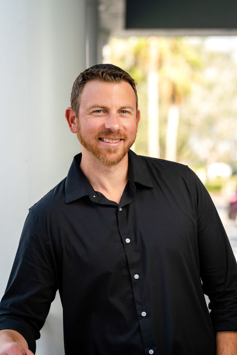 Professional wearing black button-down shirt smiling warmly at camera in business portrait.