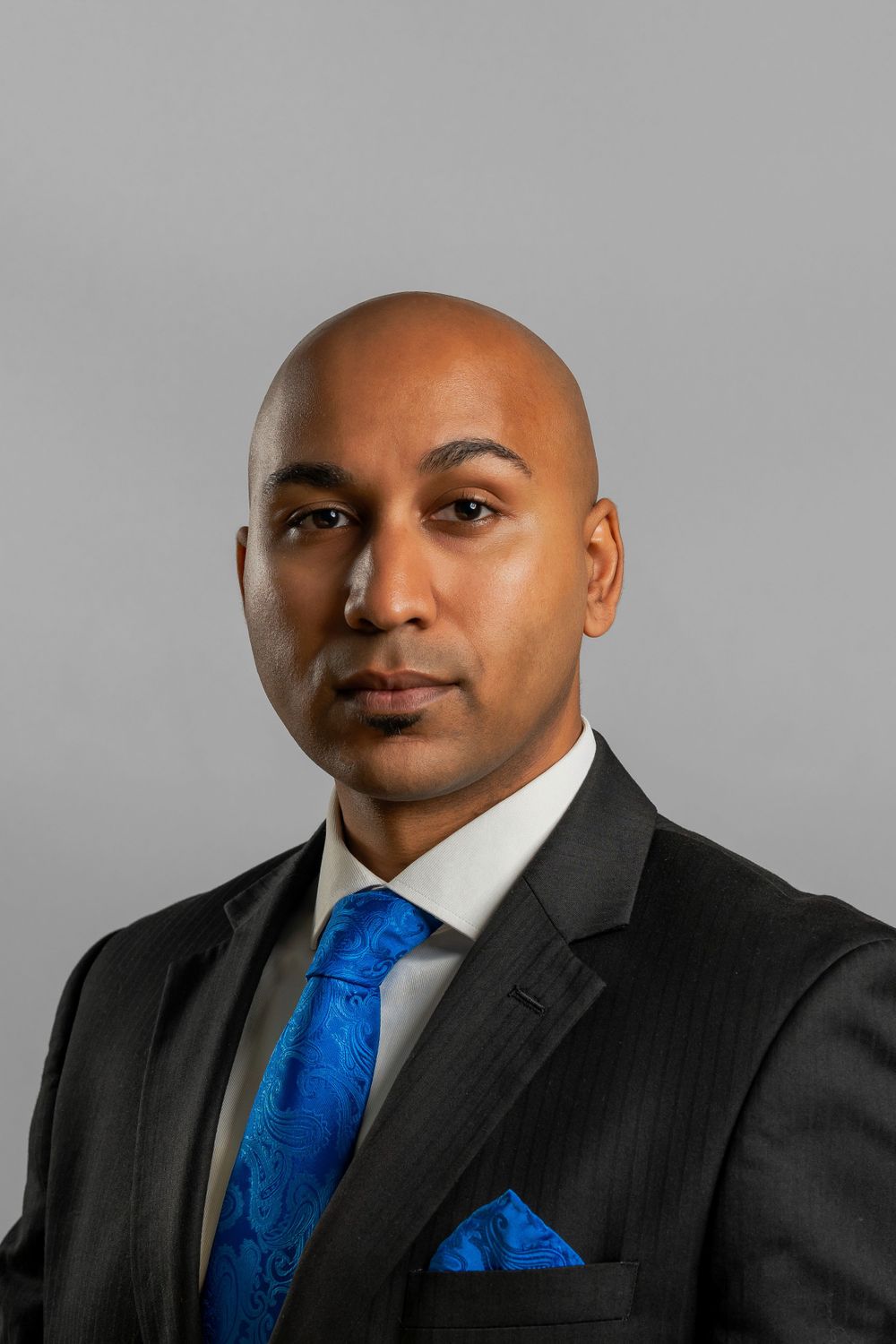 Corporate headshot in dark suit with blue tie against neutral gray background.