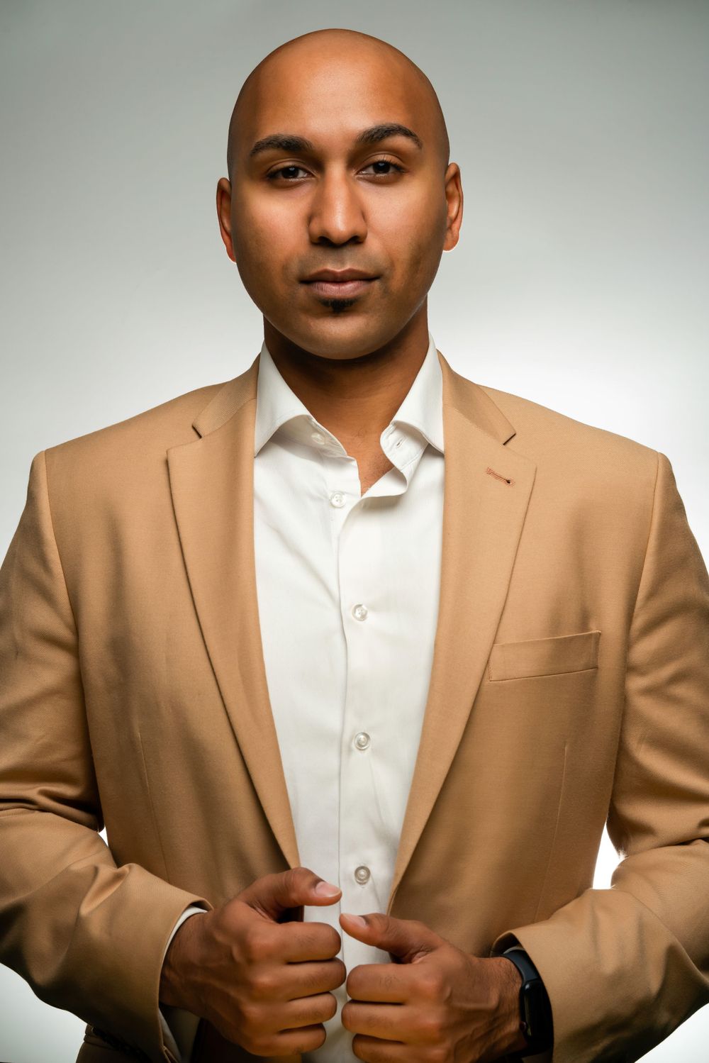 Professional studio portrait in tan suit jacket and white dress shirt against gray background.