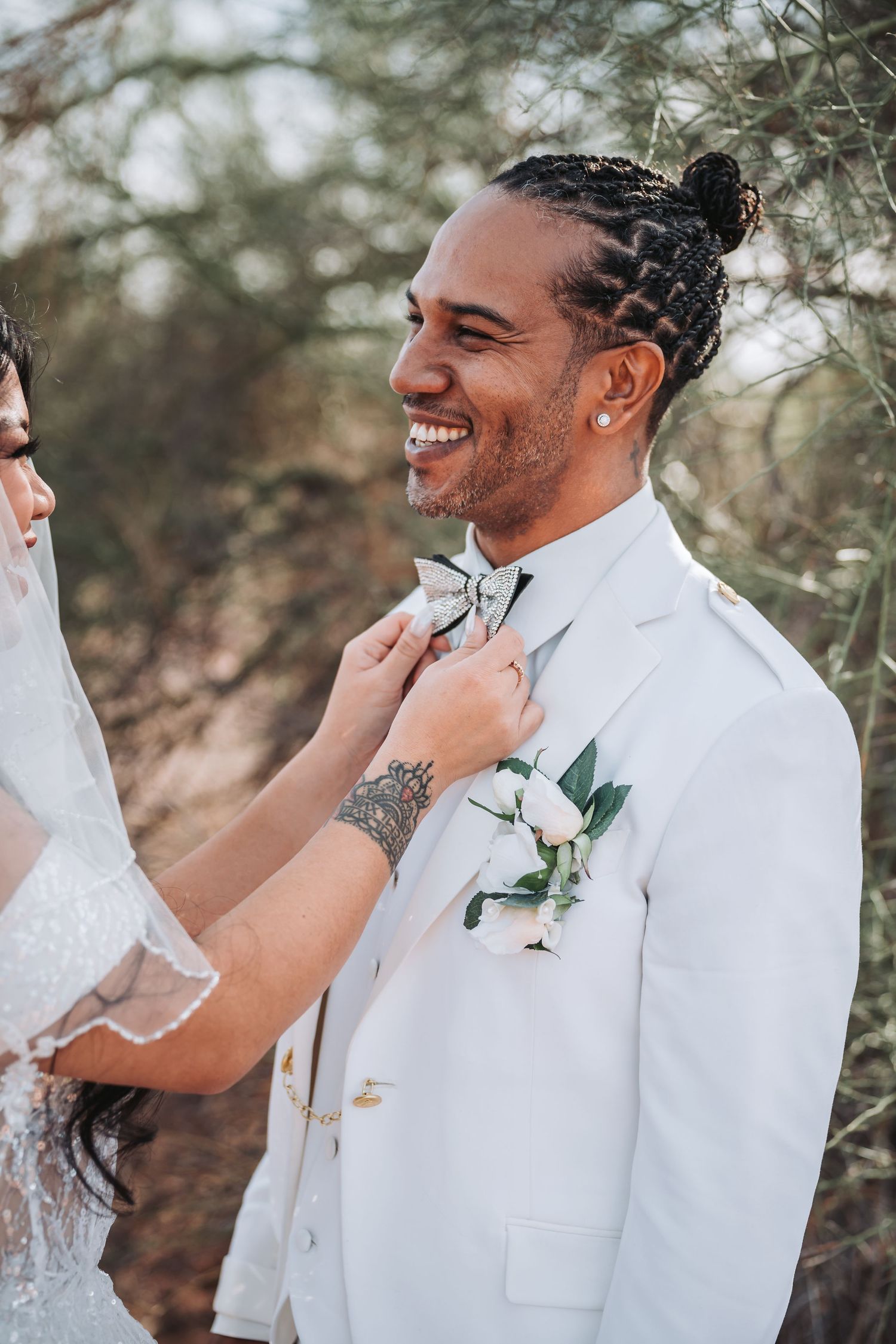 A groom in a white suit smiles while getting boutonniere adjusted during an outdoor wedding.