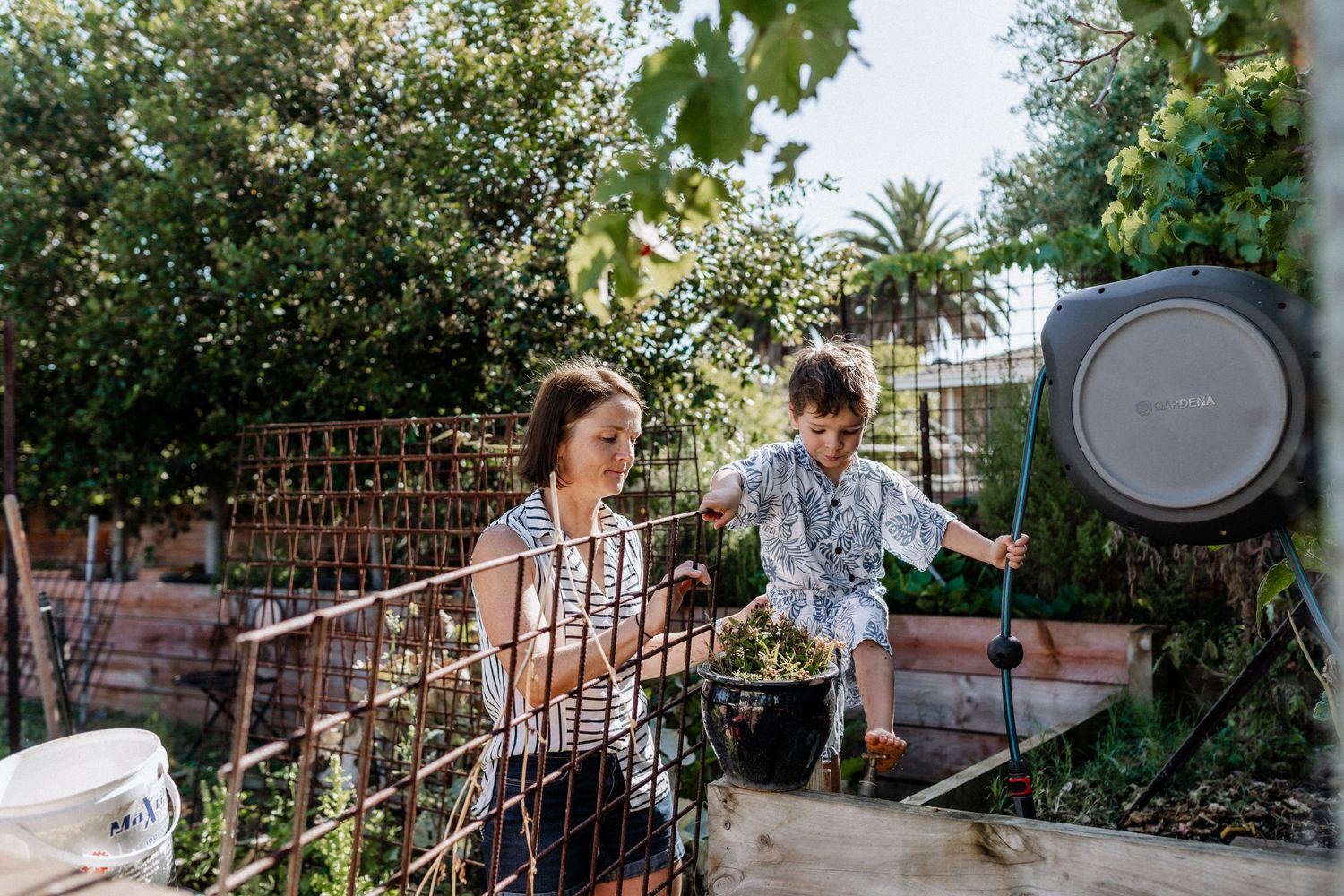 A mother holds the hand of her young son balancing on a wall in their backyard. U