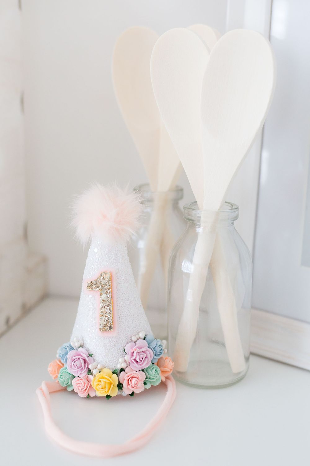A decorative first birthday party hat with pastel flowers and two white heart-shaped fans in glass bottles.