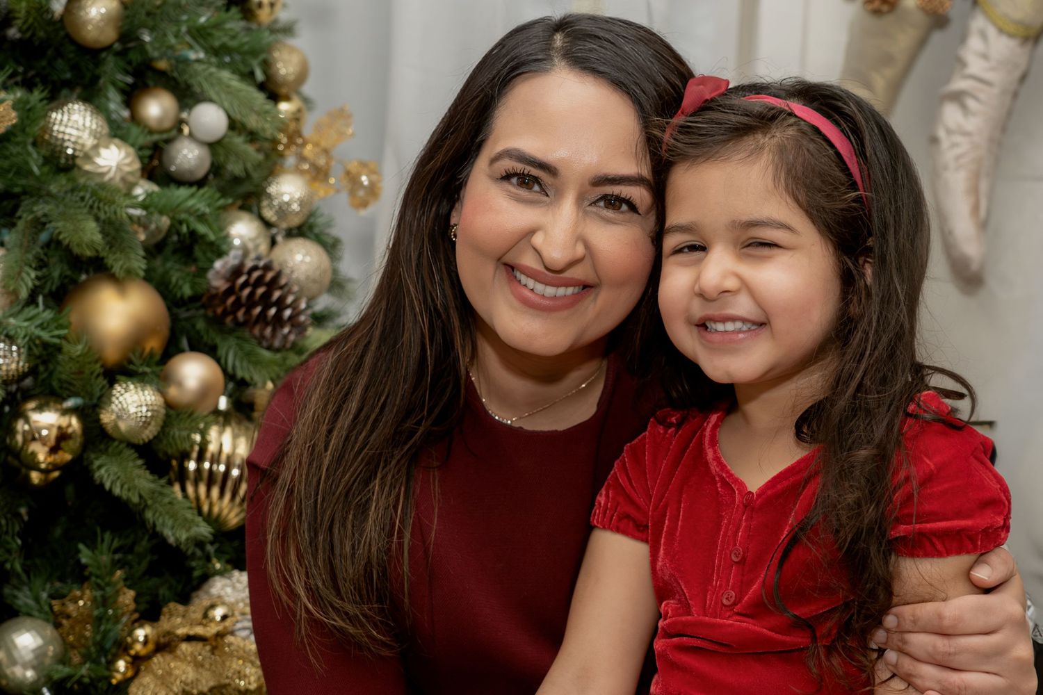 Mother and daughter in matching red outfits pose together in front of a decorated Christmas tree with gold ornaments.