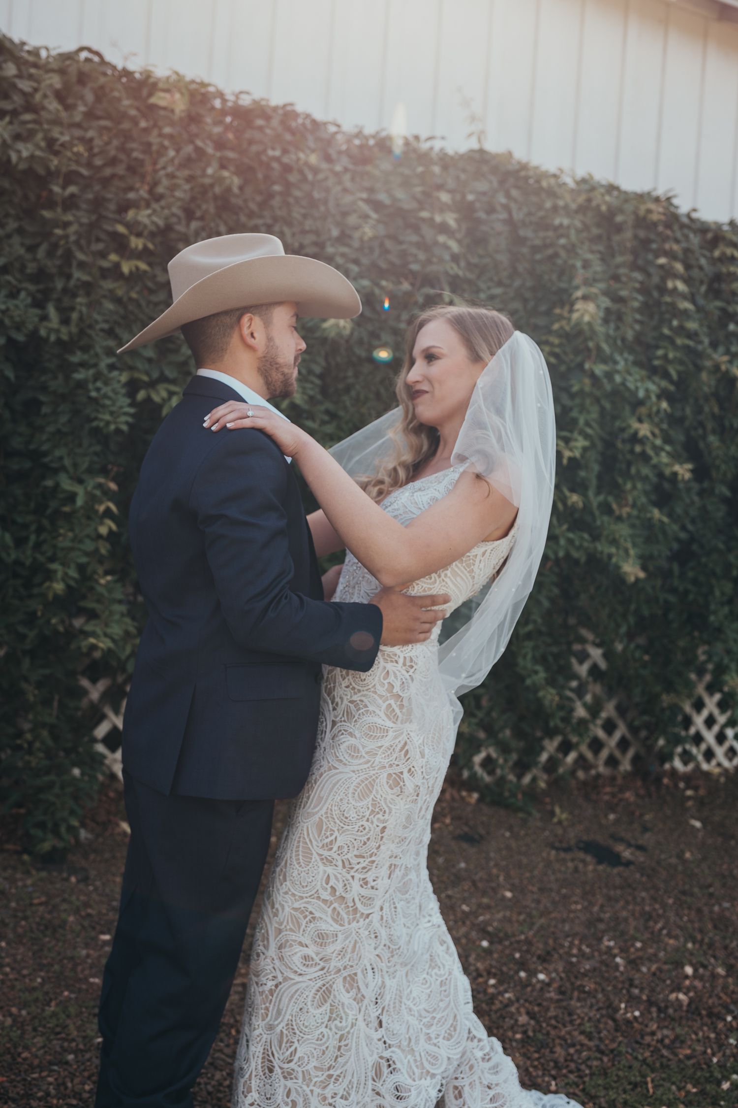 A wedding couple in formal attire embrace outside against a hedge, with the bride in lace and groom in cowboy hat.
