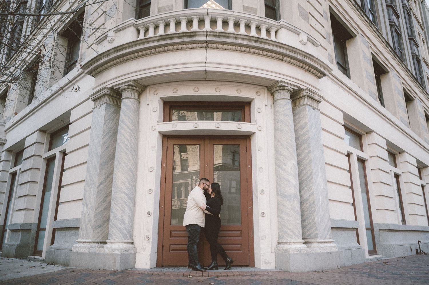 A couple embraces in front of ornate wooden doors at the corner entrance of a historic stone building.