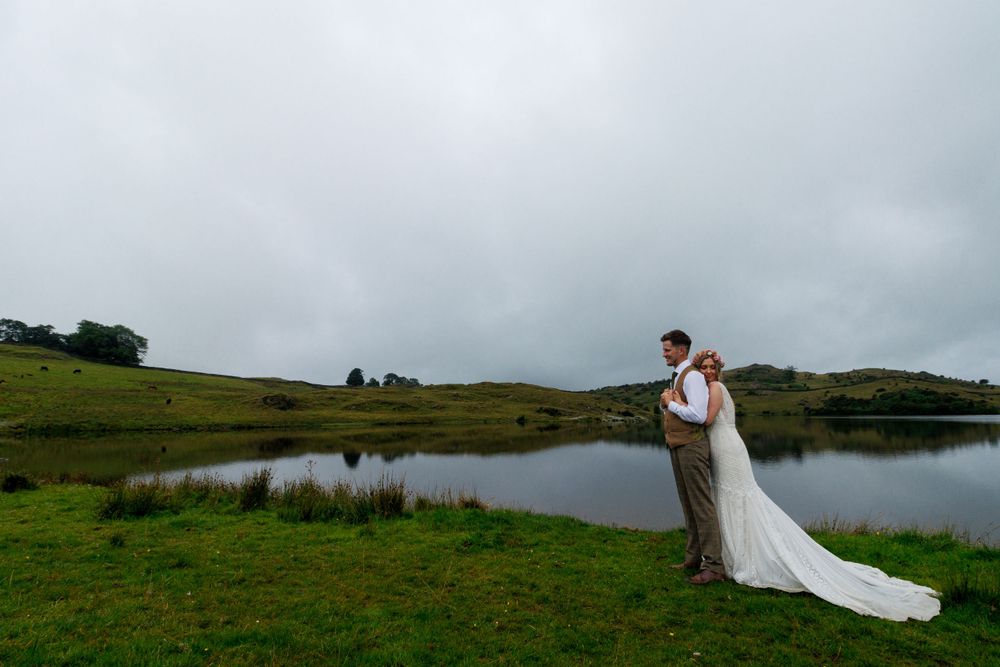 Wedding couple standing by a serene lake surrounded by rolling green hills under moody skies.