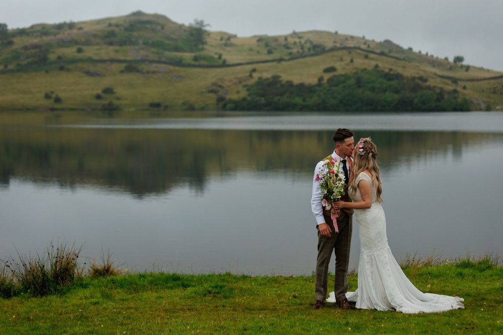 A romantic wedding moment captured by a peaceful lake with misty hills in the background.