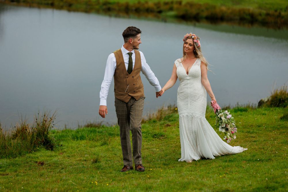 A couple walks hand in hand through green grass beside a peaceful lake during their wedding photoshoot.