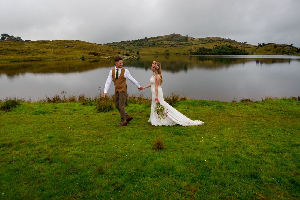 Bride and groom walk together on green grass with a serene lake and rolling hills behind them.