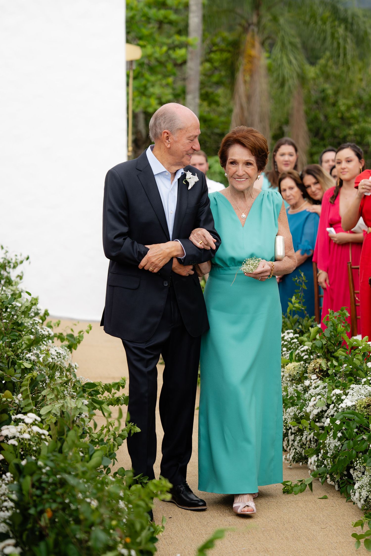 Elegant couple walks arm in arm down a garden path while wedding party in red dresses looks on from behind.