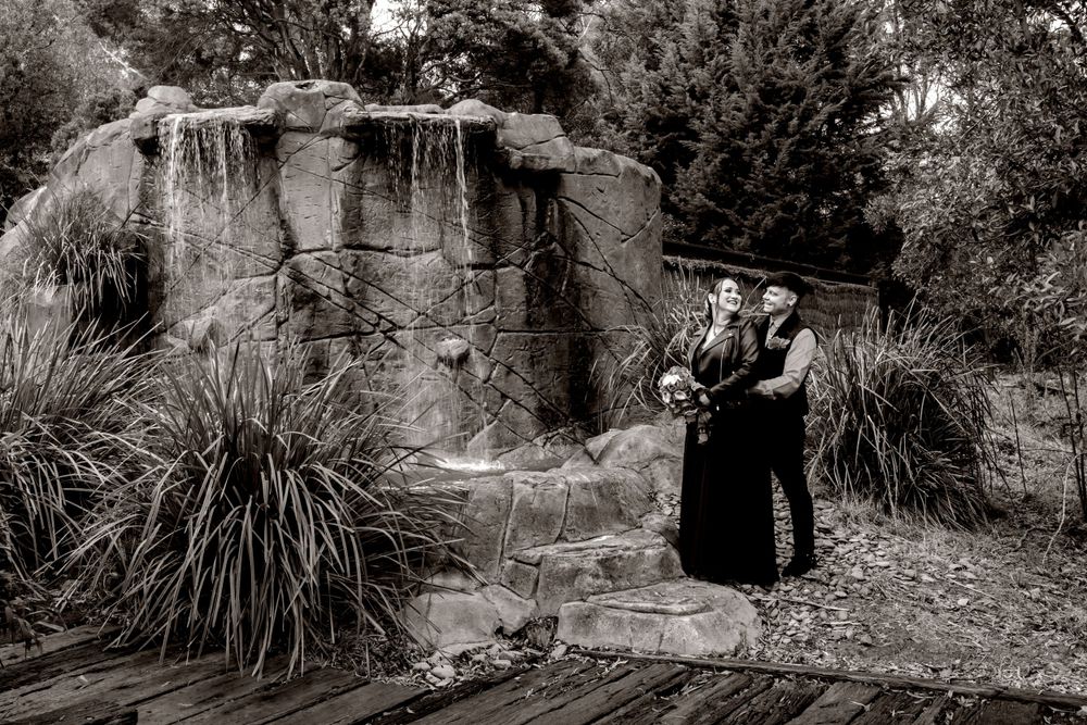 Bride in black dress and groom in black vest A black and white photo of two people standing by stone ruins surrounded by ornamental grasses.