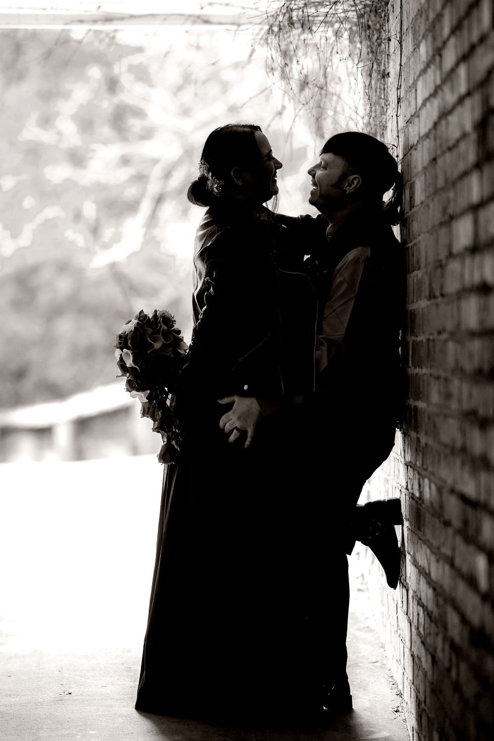 Bride in black dress and groom in black vest in Silhouetted figures against brick wall in moody black and white photo sequence.