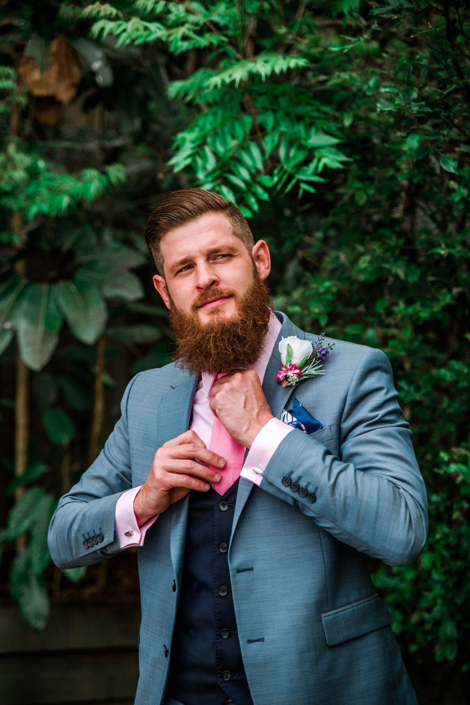 A bearded groom in a grey suit and pink tie adjusts his collar against a leafy backdrop.
