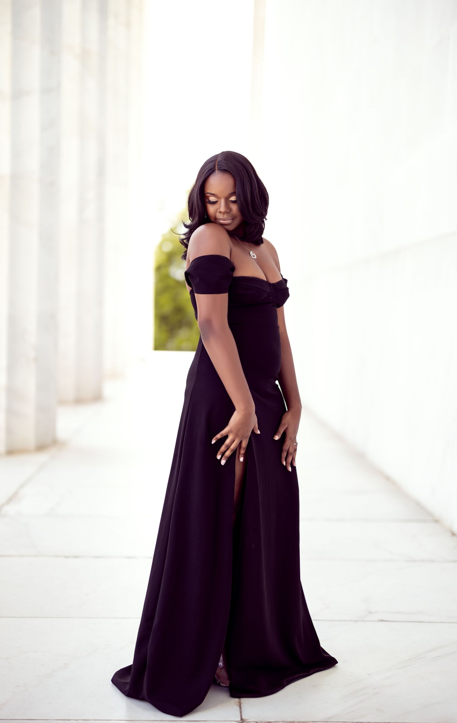 A woman in an elegant black off-shoulder gown poses in a bright white hallway.