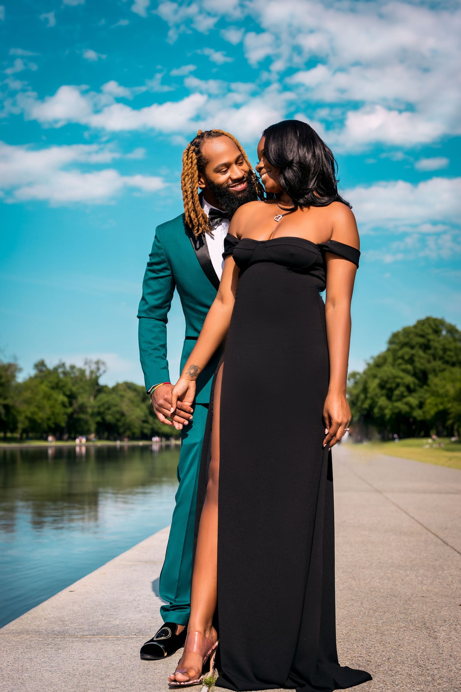 A romantic couple embraces on a pathway next to a lake under blue skies.