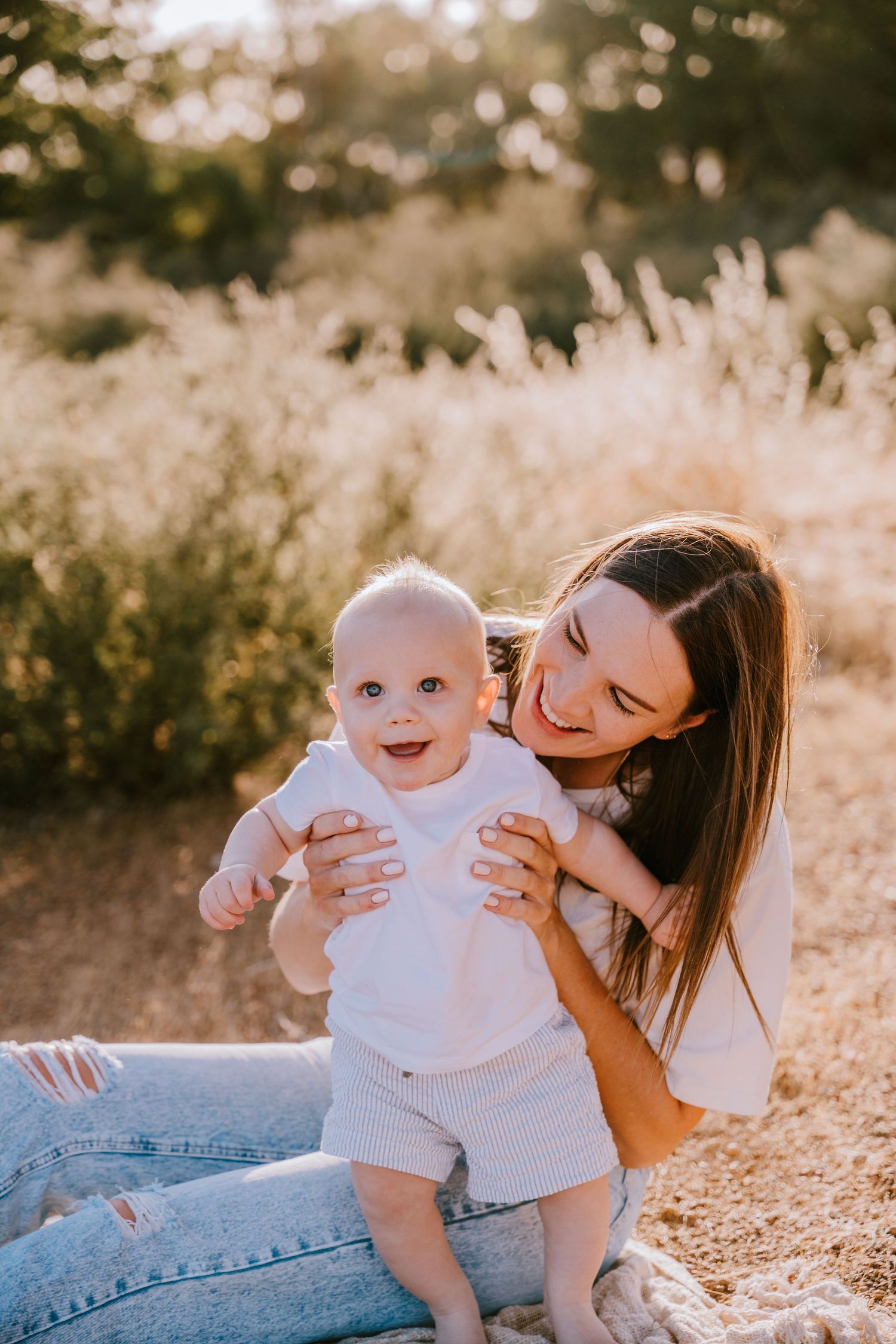 A heartwarming outdoor portrait of a baby standing with support amid sun-drenched grass and foliage.