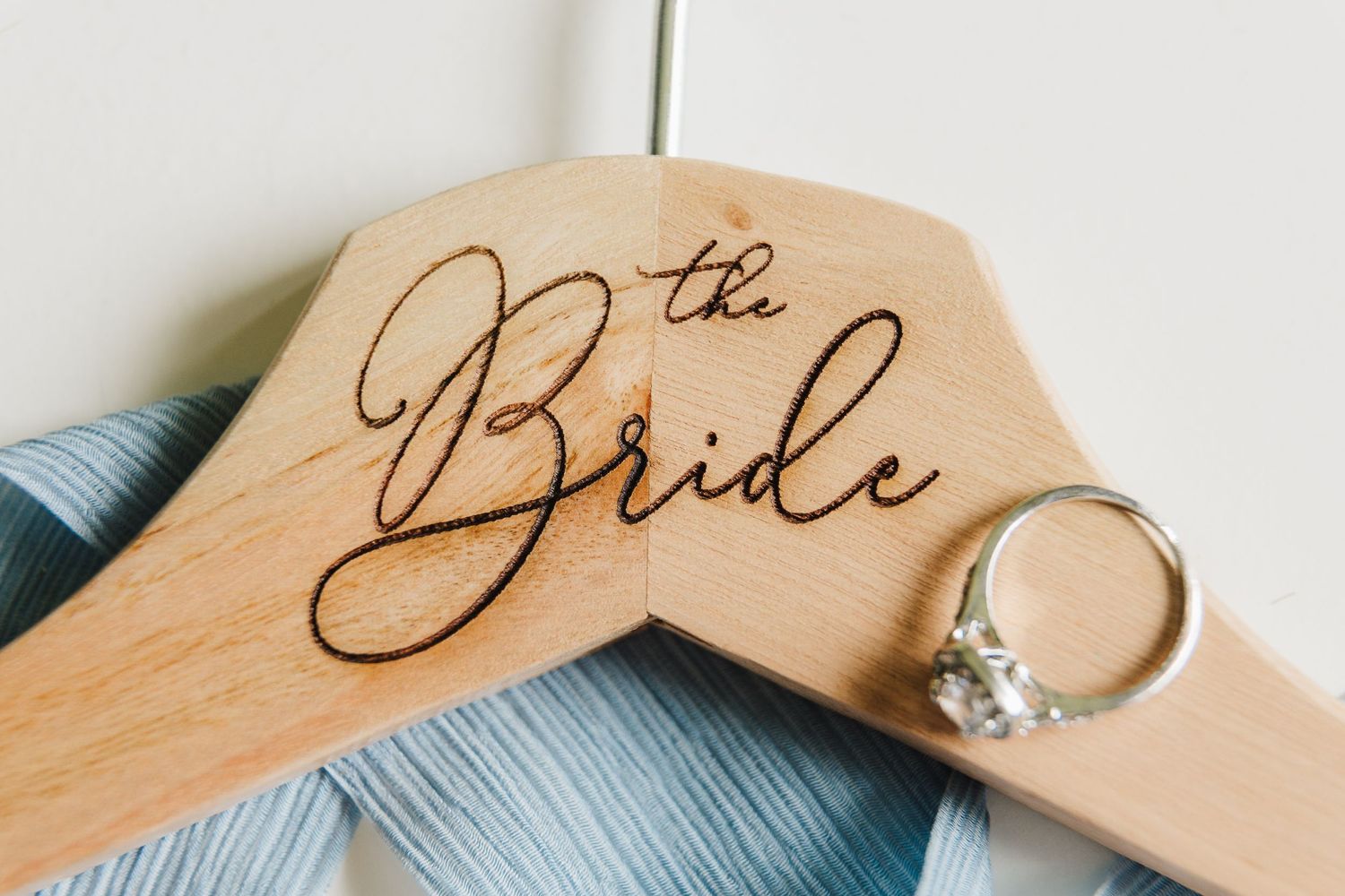 Wooden hanger engraved with 'The Bride' text and engagement ring against blue fabric.