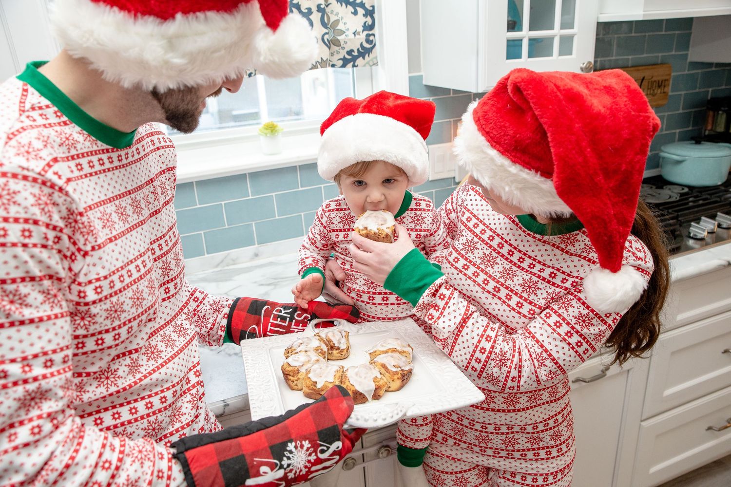 mom and dad and toddler Christmas pajamas Santa hats eating cinnamon rolls in kitchen Christmas photos