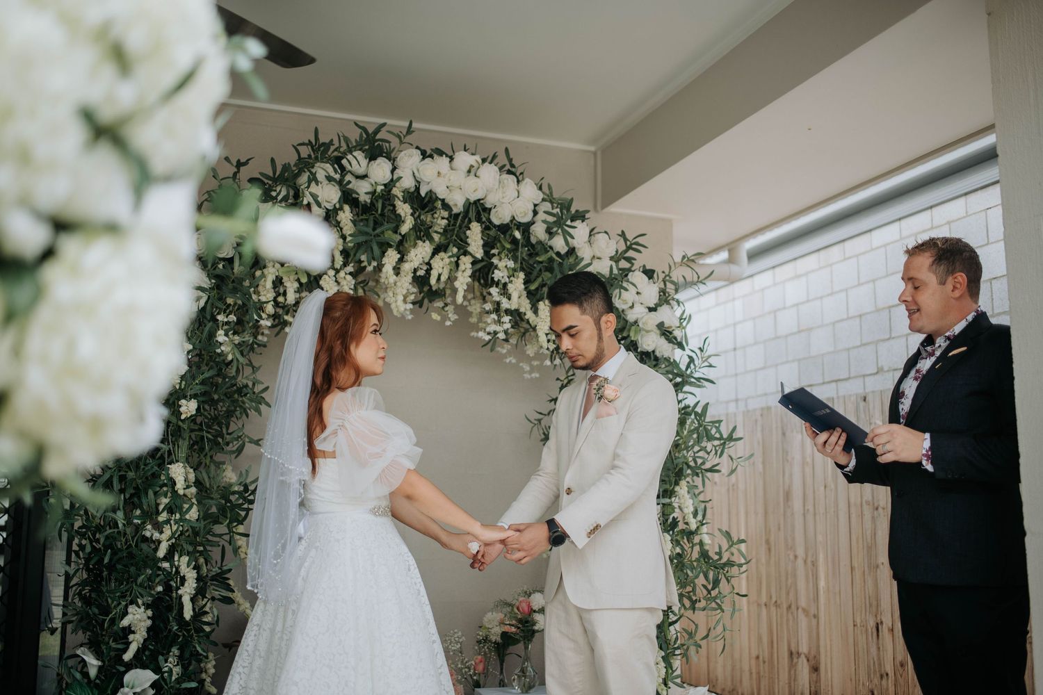 Wedding ceremony under a white floral arch with greenery indoors.