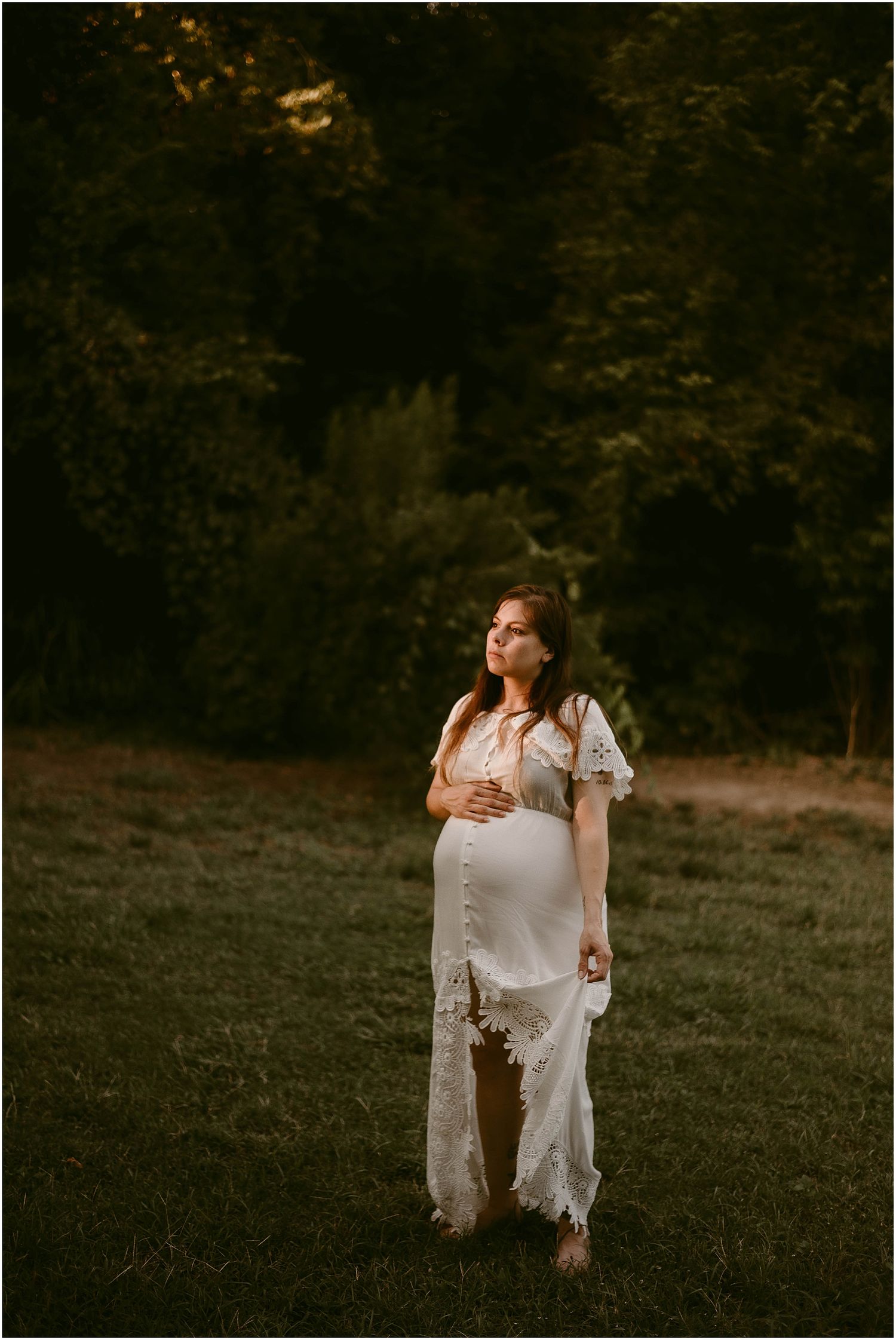 Atmospheric maternity portrait in a white lace gown against a moody forest backdrop.