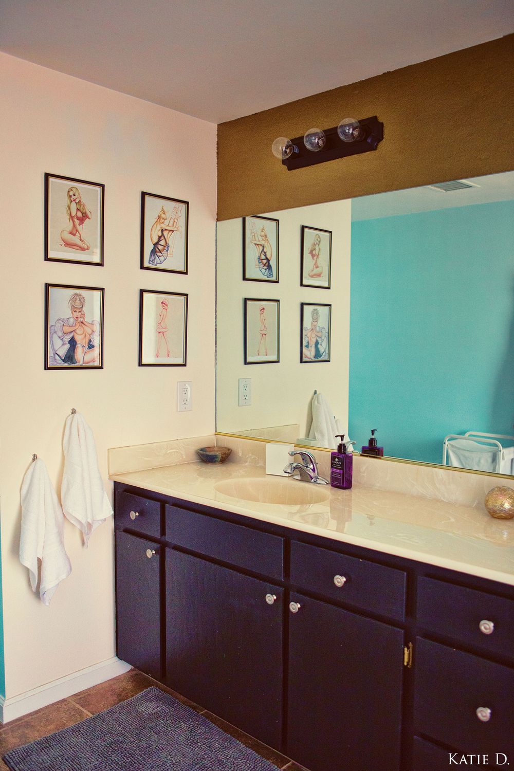 Modern bathroom vanity with dark cabinets, large mirror, and vintage pin-up art prints on beige walls.