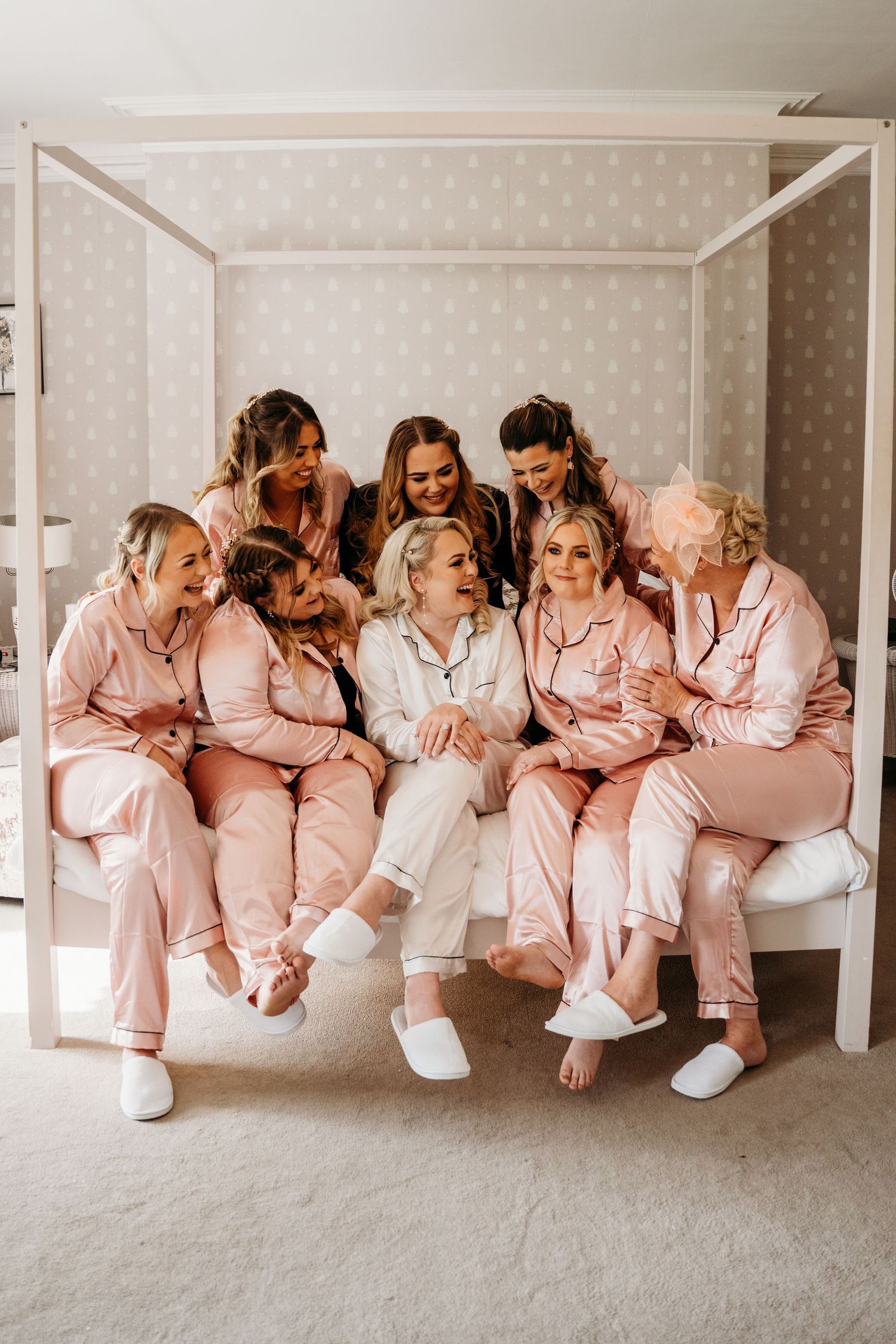 Group in matching pink satin pajamas sharing laughter on white canopy bed.