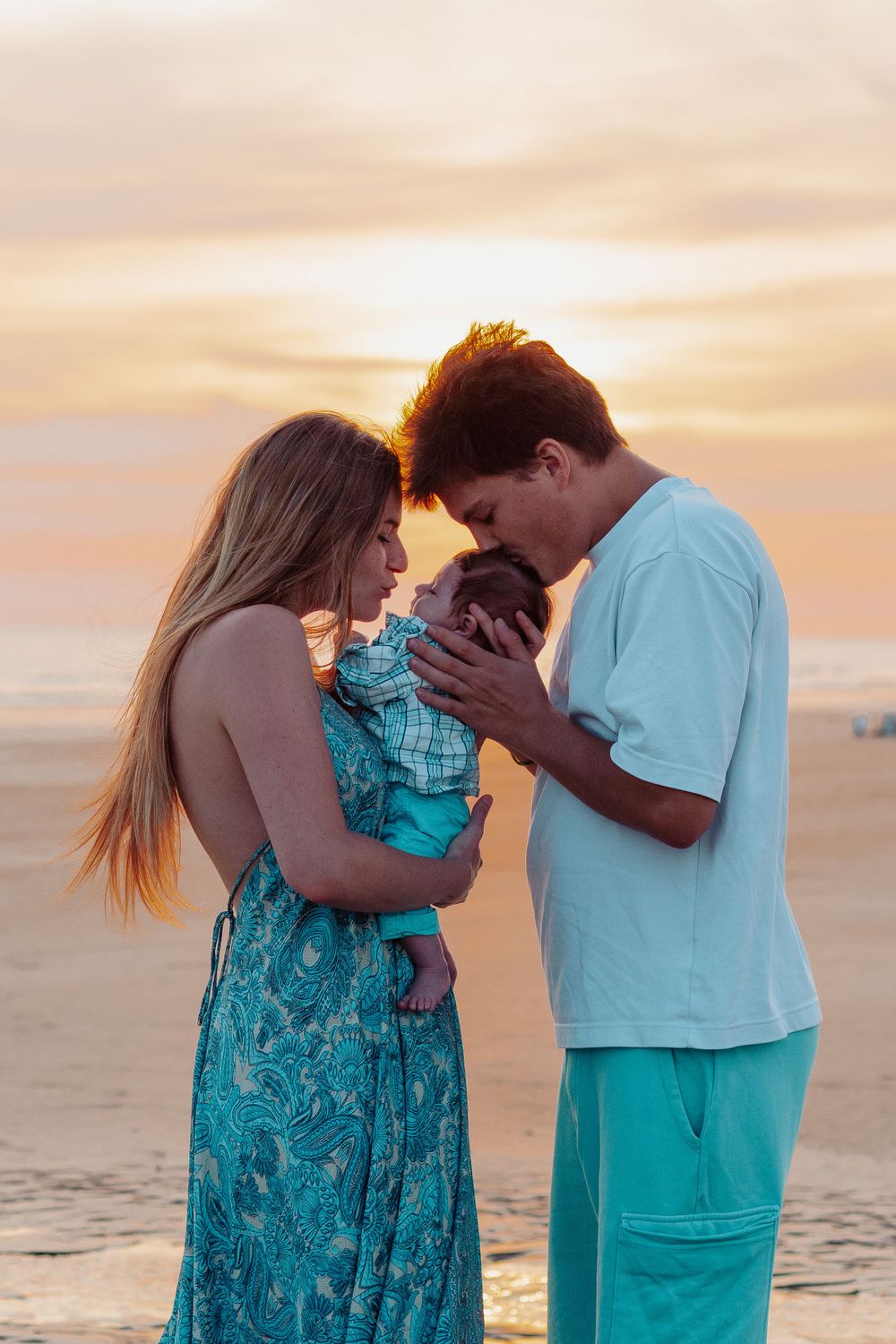 Family shares intimate moment at sunset on beach wearing coordinating turquoise and white outfits.