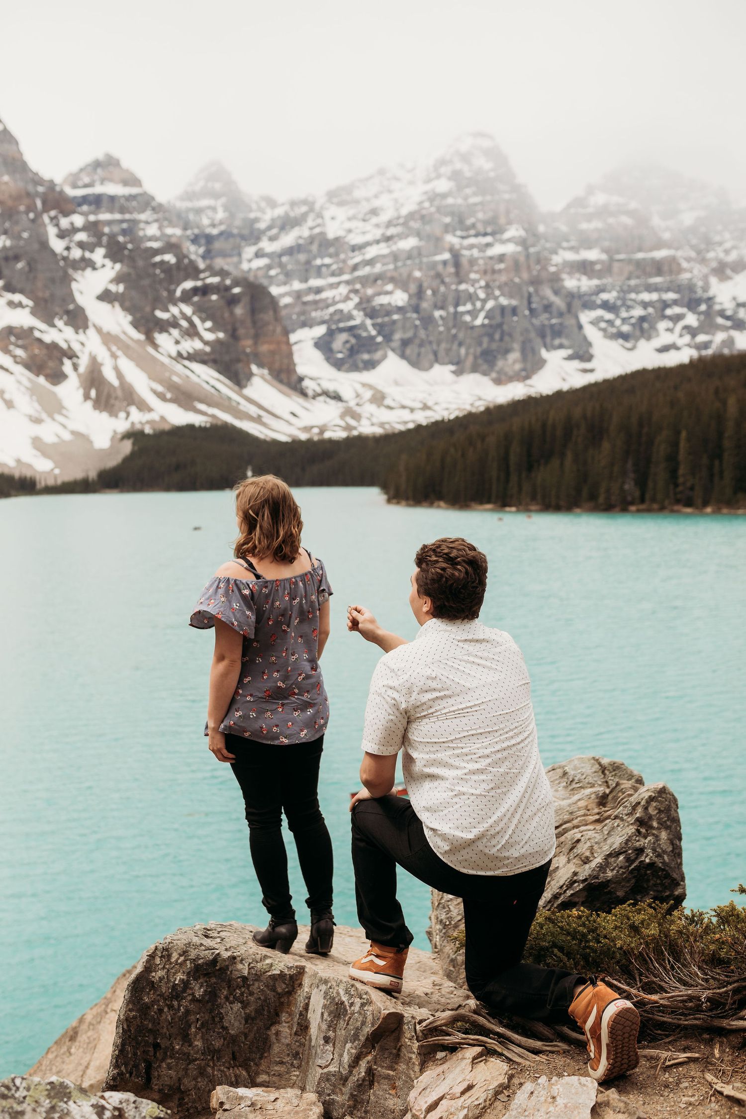 Marriage proposal scene at scenic mountain lake overlook with snow-capped peaks.