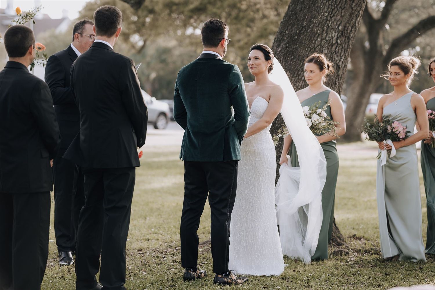 Outdoor wedding ceremony with bridal party in black suits and sage green dresses.
