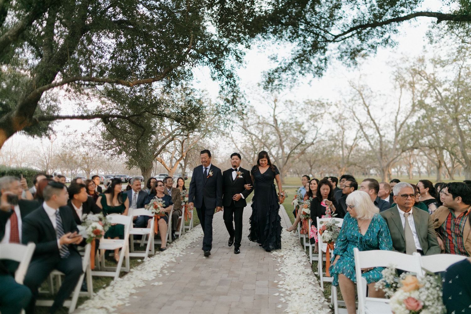 Wedding ceremony processional on a stone path lined with guests in white chairs under trees.