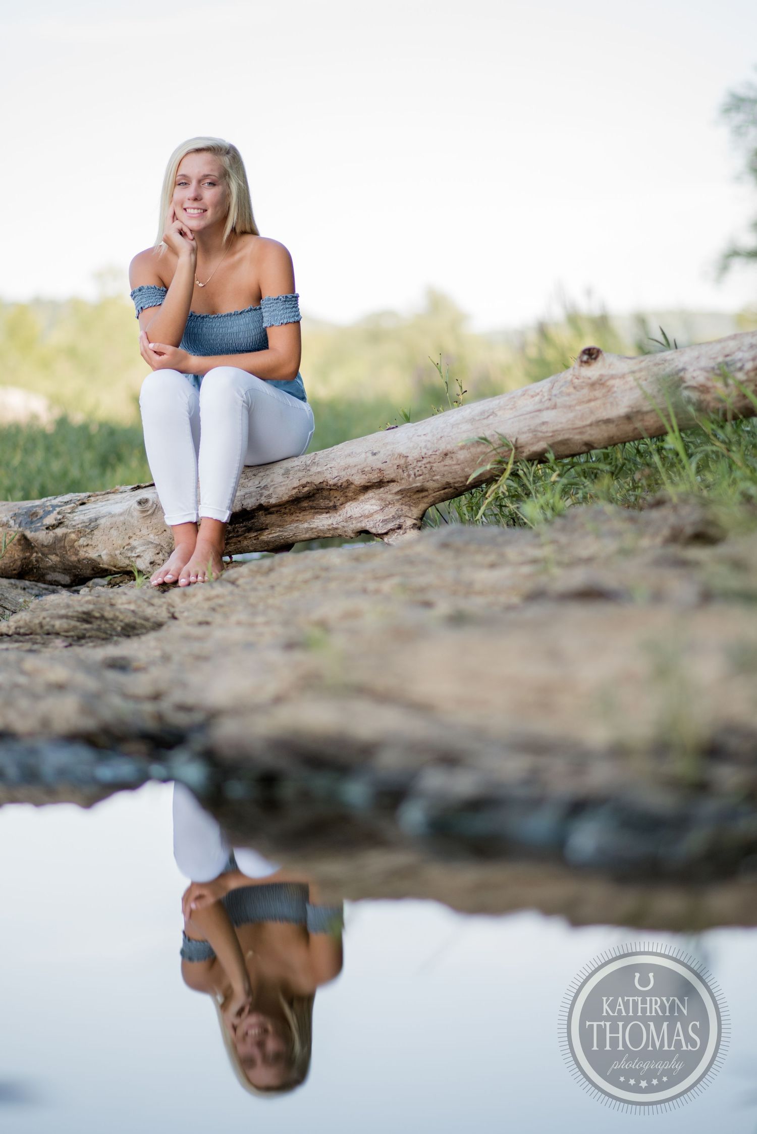 Woman in off-shoulder top sits on a log by a reflective pond in an outdoor setting.