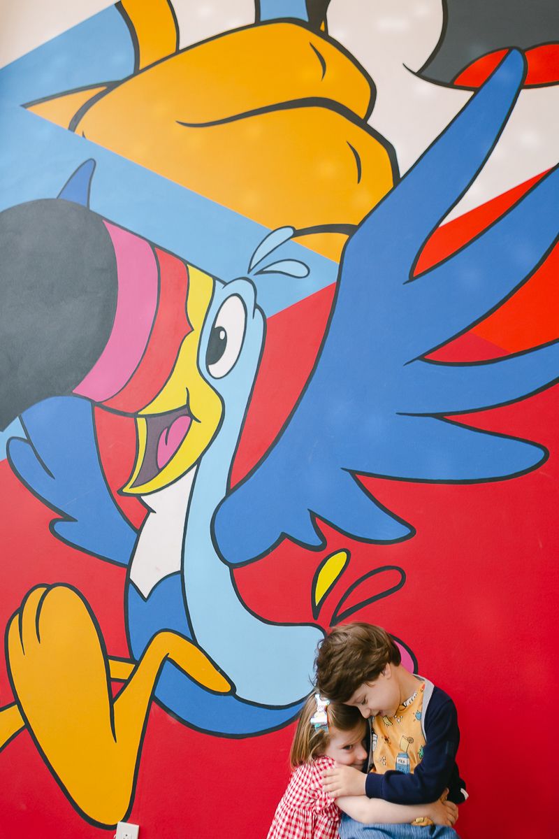 an open view of siblings hugging with a Kellogg's tucan mural in the background photographed by Paula Ortiz NY Family