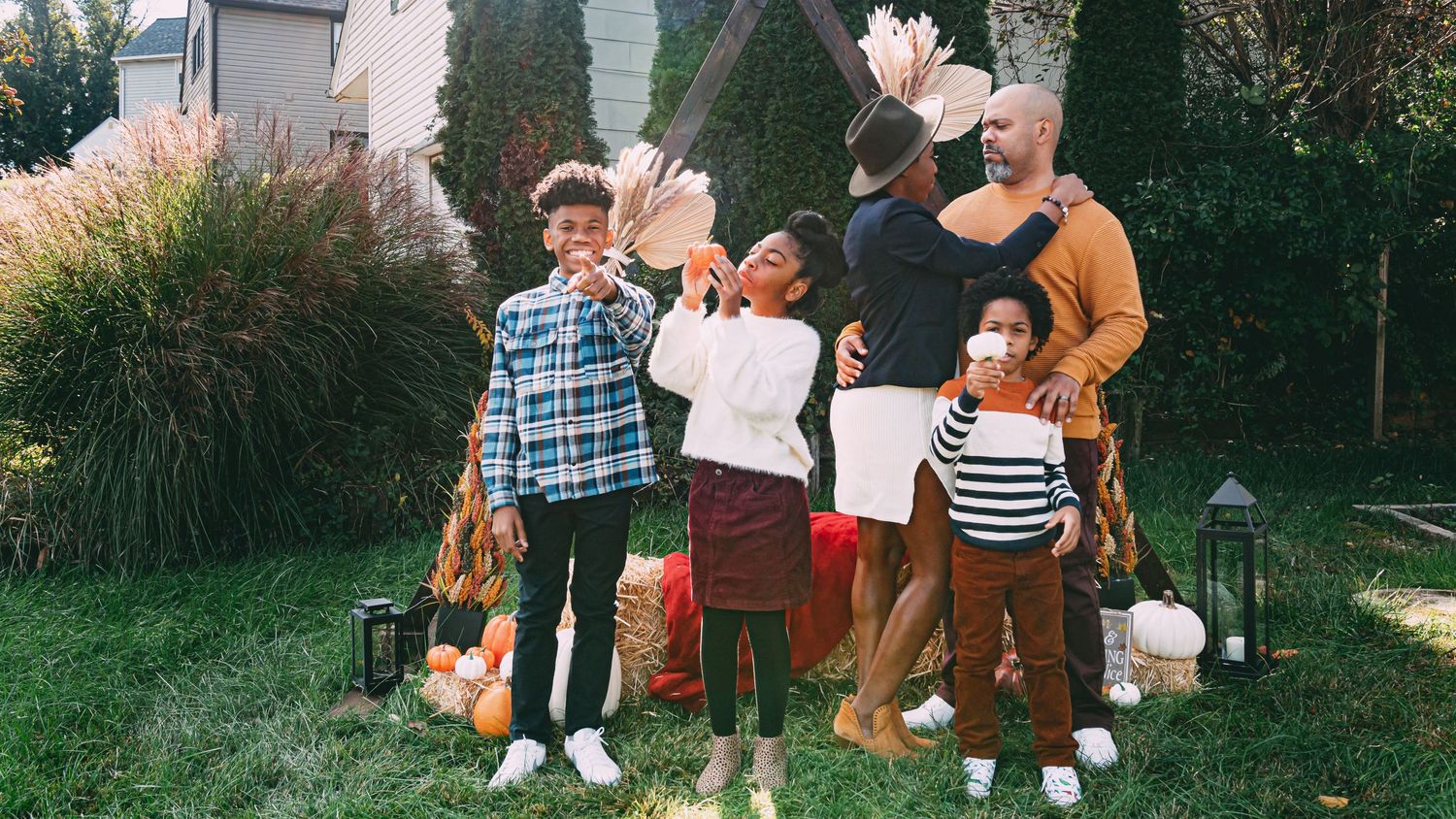 Playful family of five laughs and poses with dried pampas grass in decorated autumn backyard setting.
