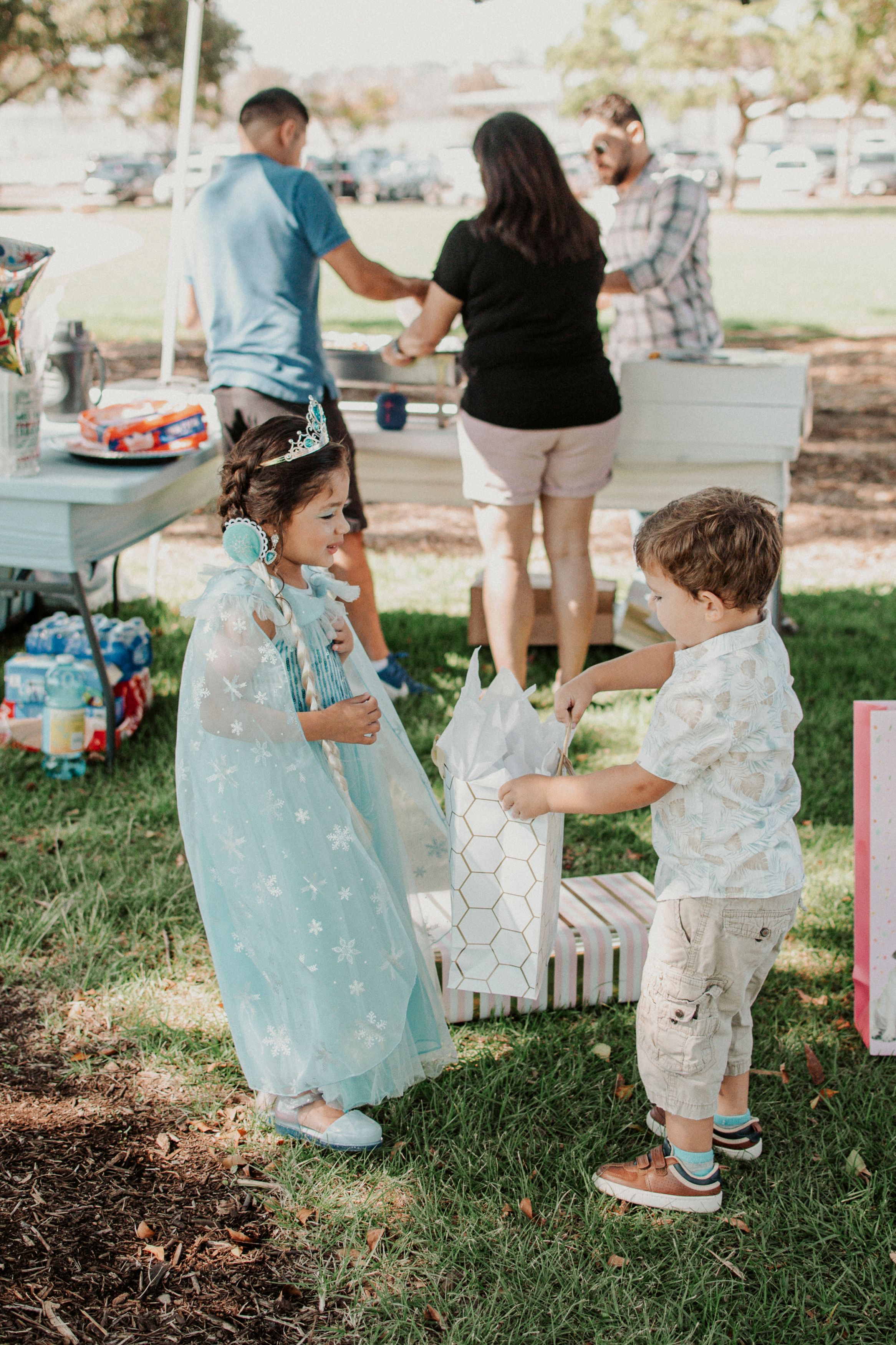 Kids in fancy clothing gather around decorative boxes on grass at an event.