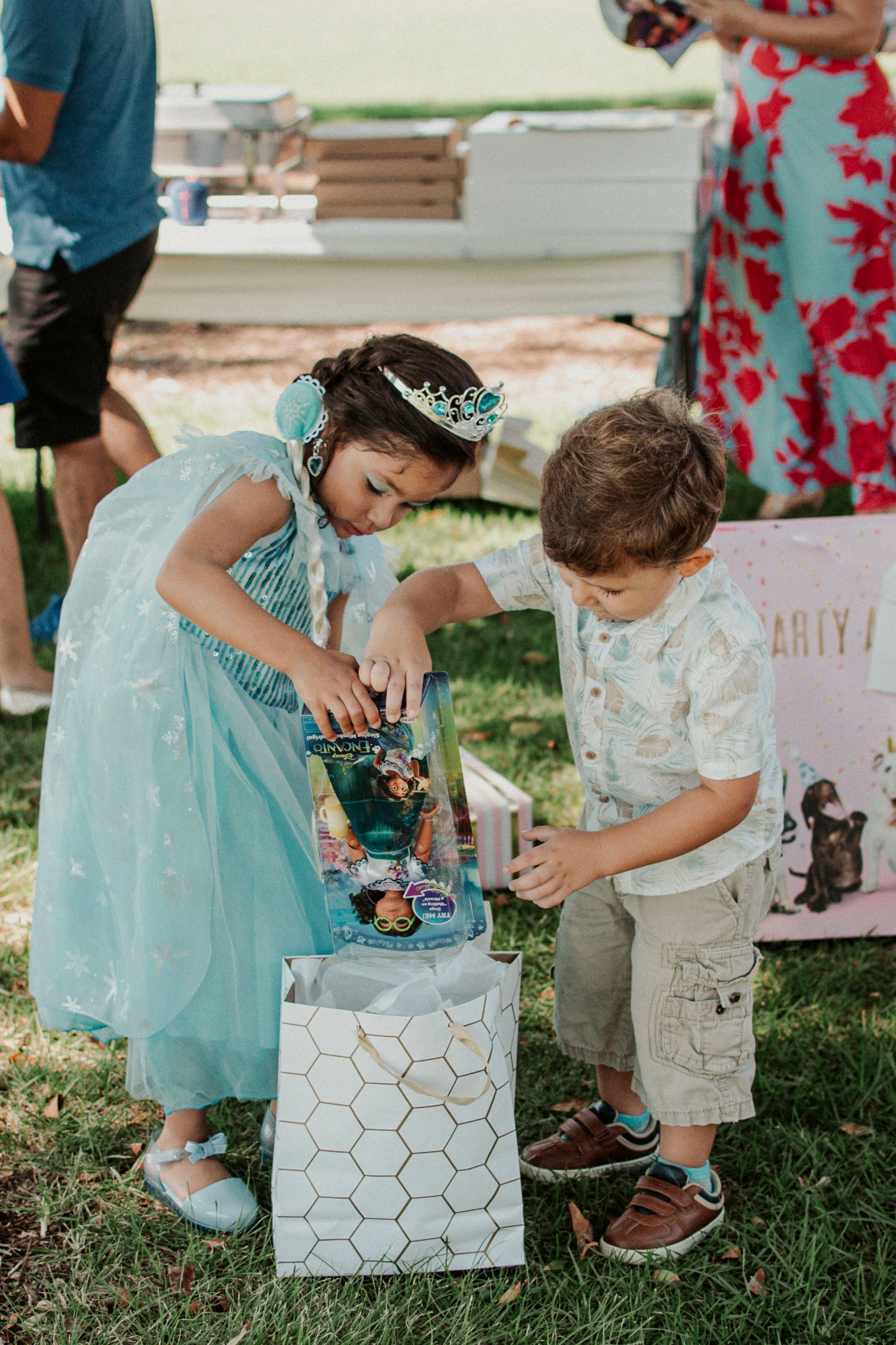 Children in formal attire play with gift boxes during an outdoor party.