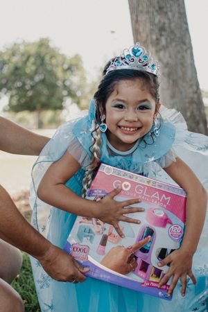 A child in a tiara and blue dress shows excitement while holding gift boxes outdoors.