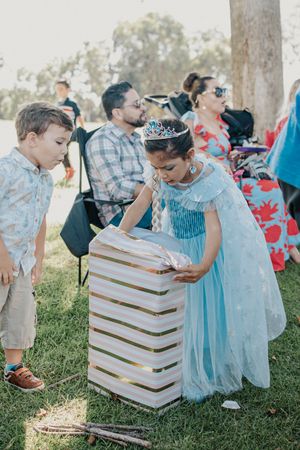 Child in blue dress opening presents next to a striped gift box at outdoor party.