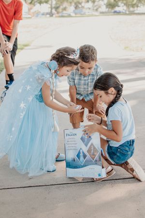 Children examining package together while wearing dress-up clothes outdoors.