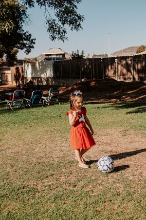 Child in orange dress standing next to soccer ball on grass field.