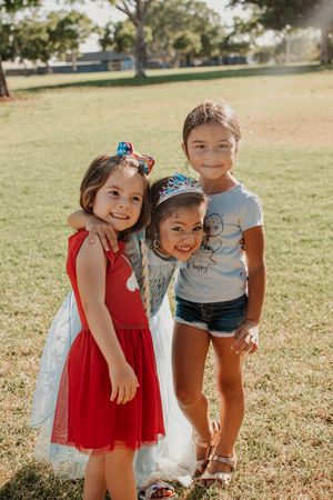 Three children in colorful dresses posing together on sunny grass field.