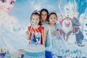 Three small children pose in front of a Frozen-themed party backdrop with Elsa and other characters.