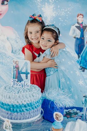 Two young children in festive dresses embrace next to a blue birthday cake with Frozen-themed decorations.