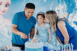 Parents and child stand by an ornate blue birthday cake with Frozen movie backdrop.