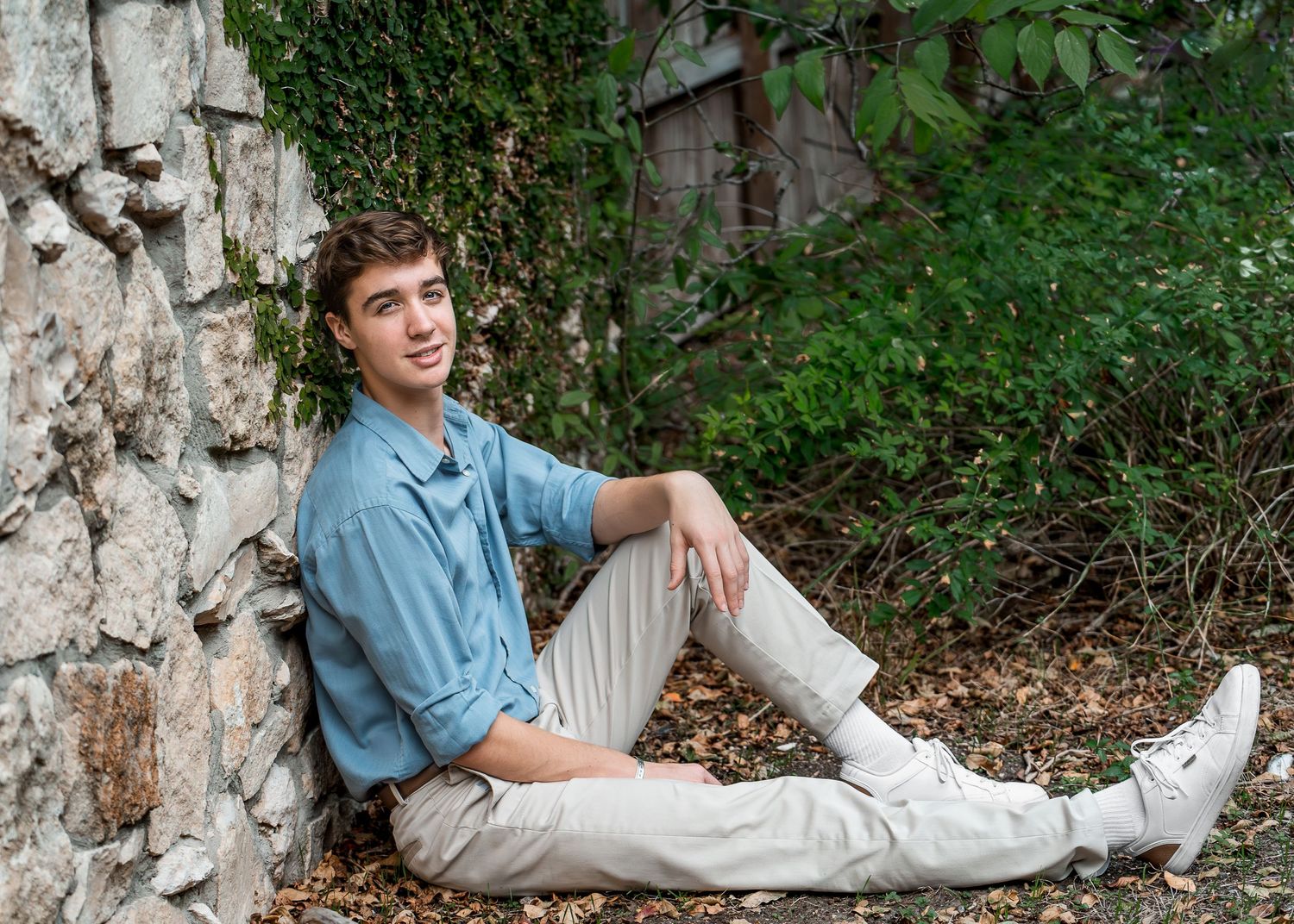 high school senior boy in blue shirt leaning against a stone wall in Landa Gardens at Landa Library